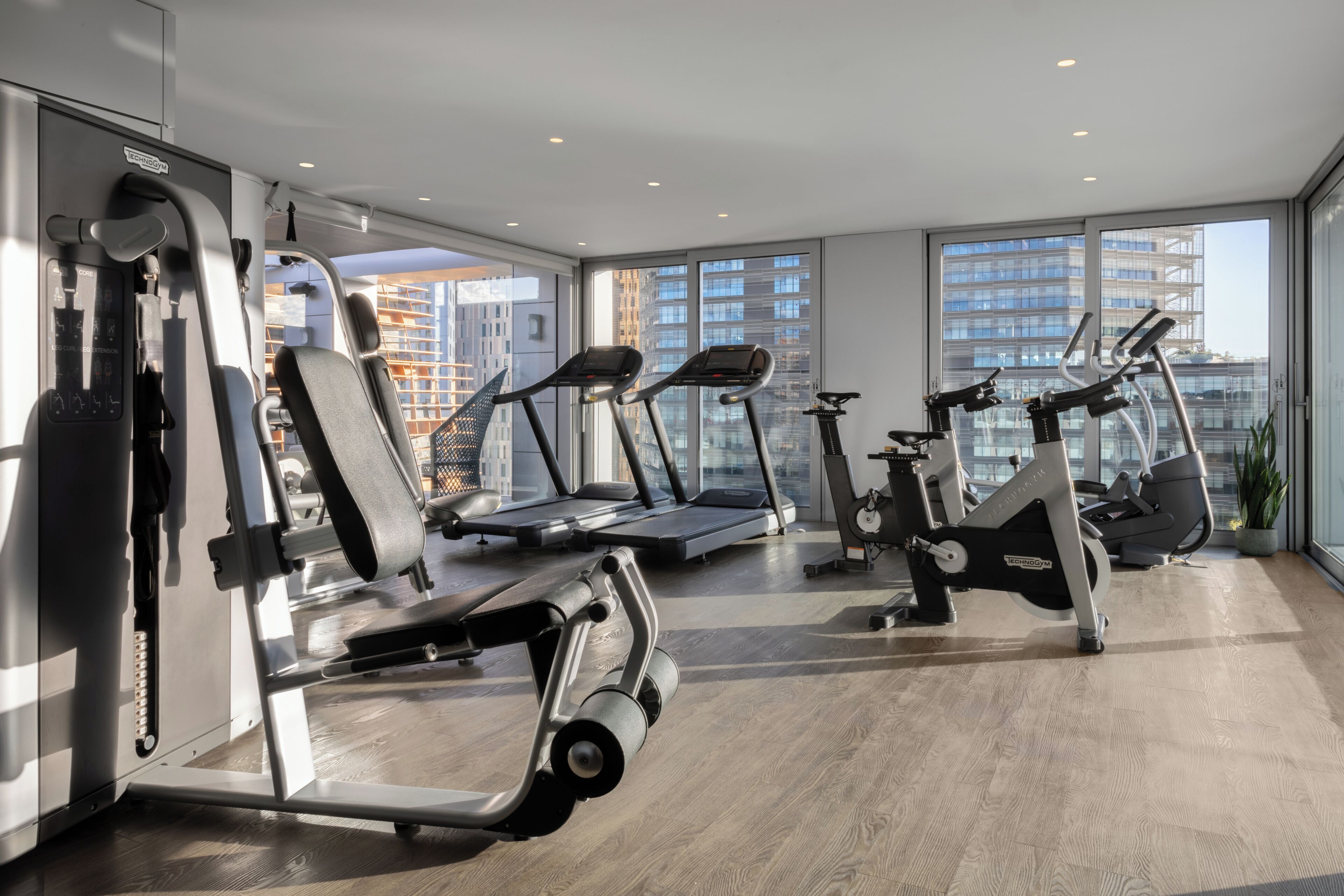A modern gym with cardio machines, weight equipment, and large windows offering city views.