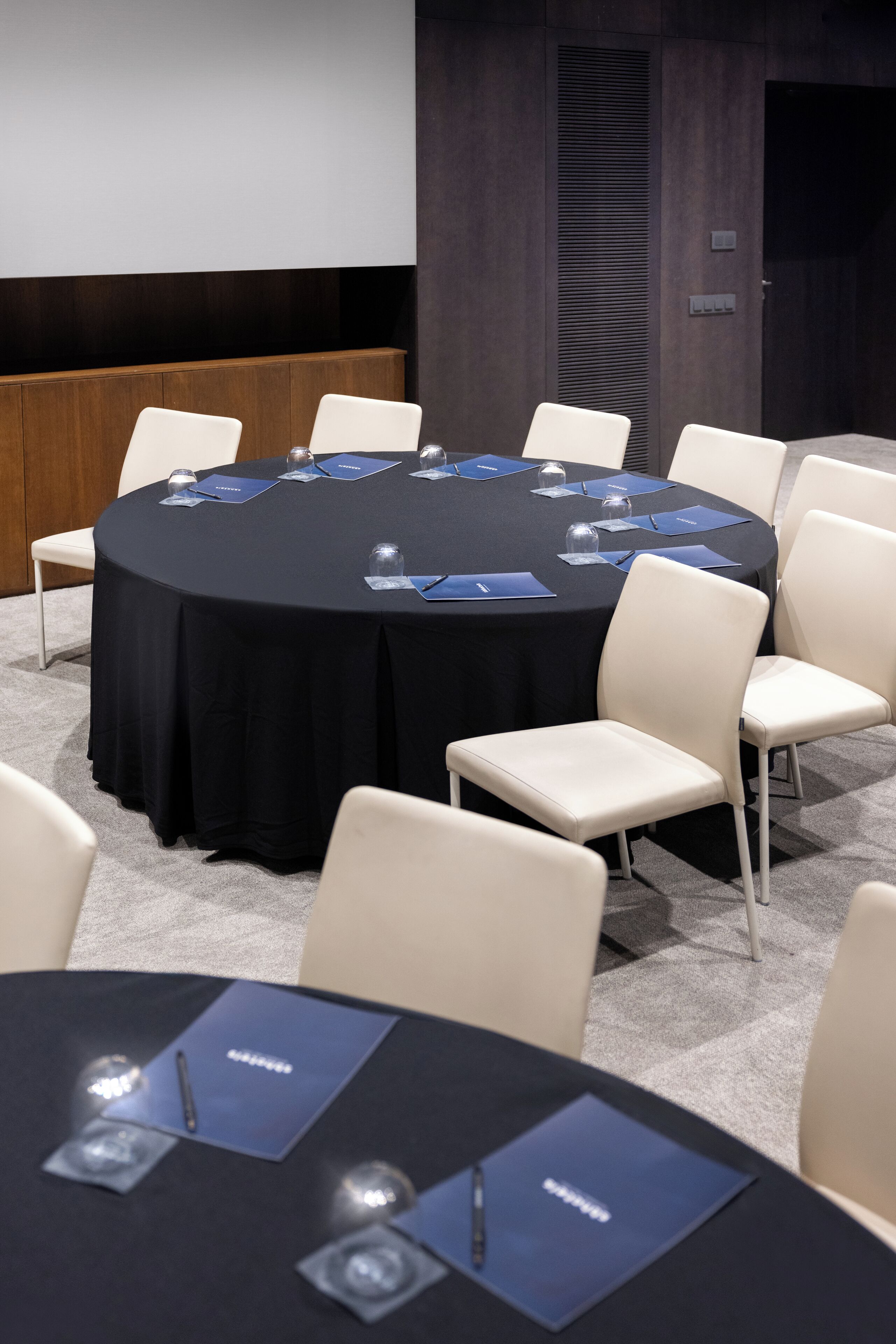 Een vergaderzaal met een ronde tafel, witte stoelen en een projectiescherm.