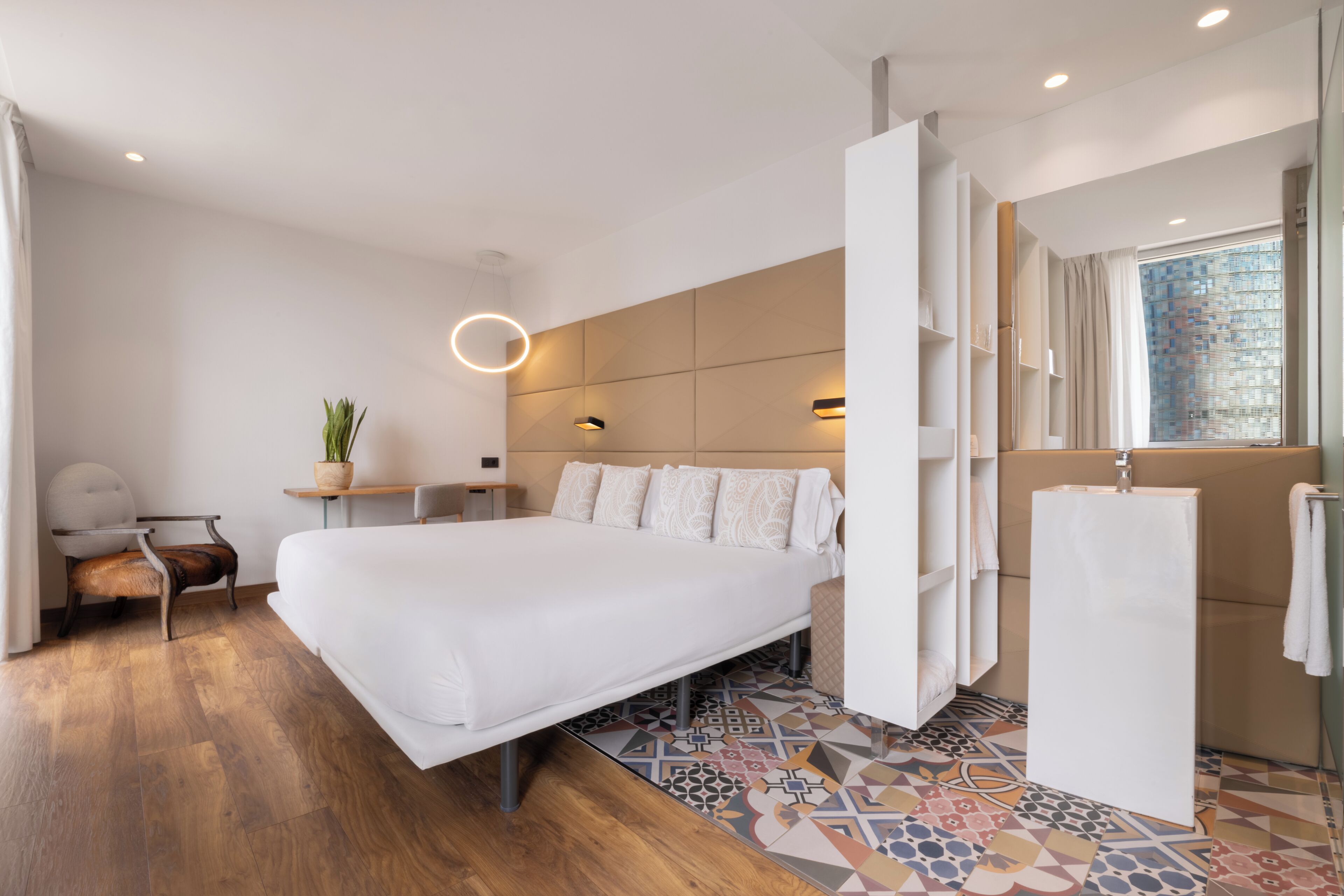 A modern hotel room with a large bed, a desk, a chair, a potted plant, a window with a view, and a bathroom area.