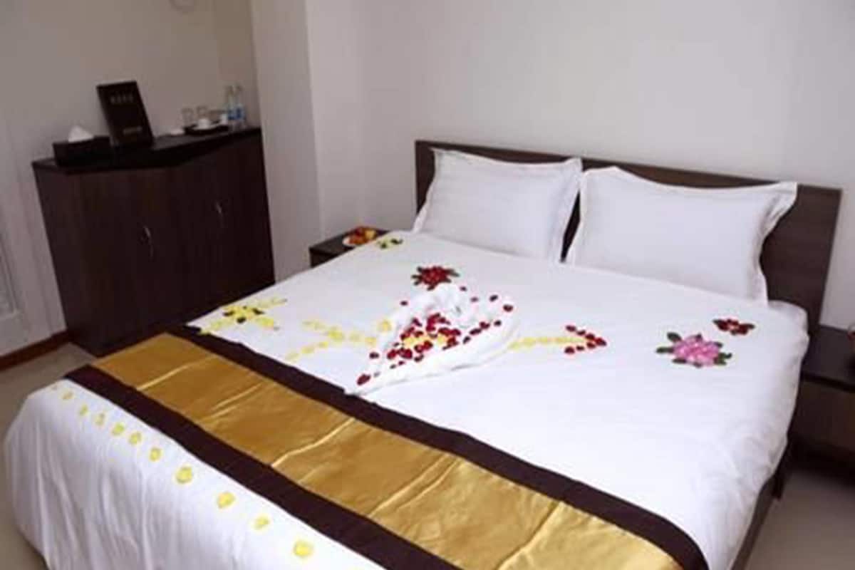 Una cama bien tendida con una colcha blanca y dorada, un cabecero de madera y una mesita de noche con una lámpara.