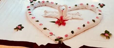 A bed with a heart-shaped towel arrangement, a purple pillow, and a decorative item on the bedspread.