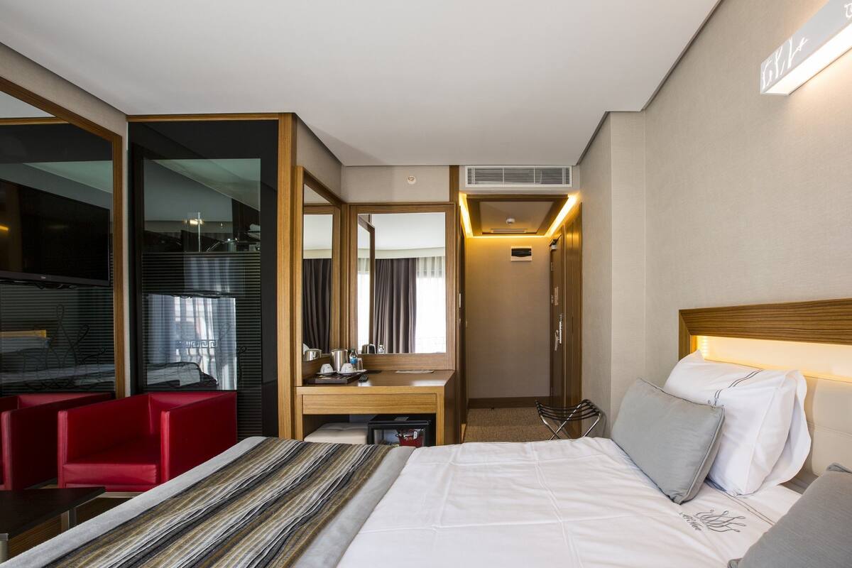 A hotel room with a bed, a desk, a red chair, a mirror, and a view of the outside.