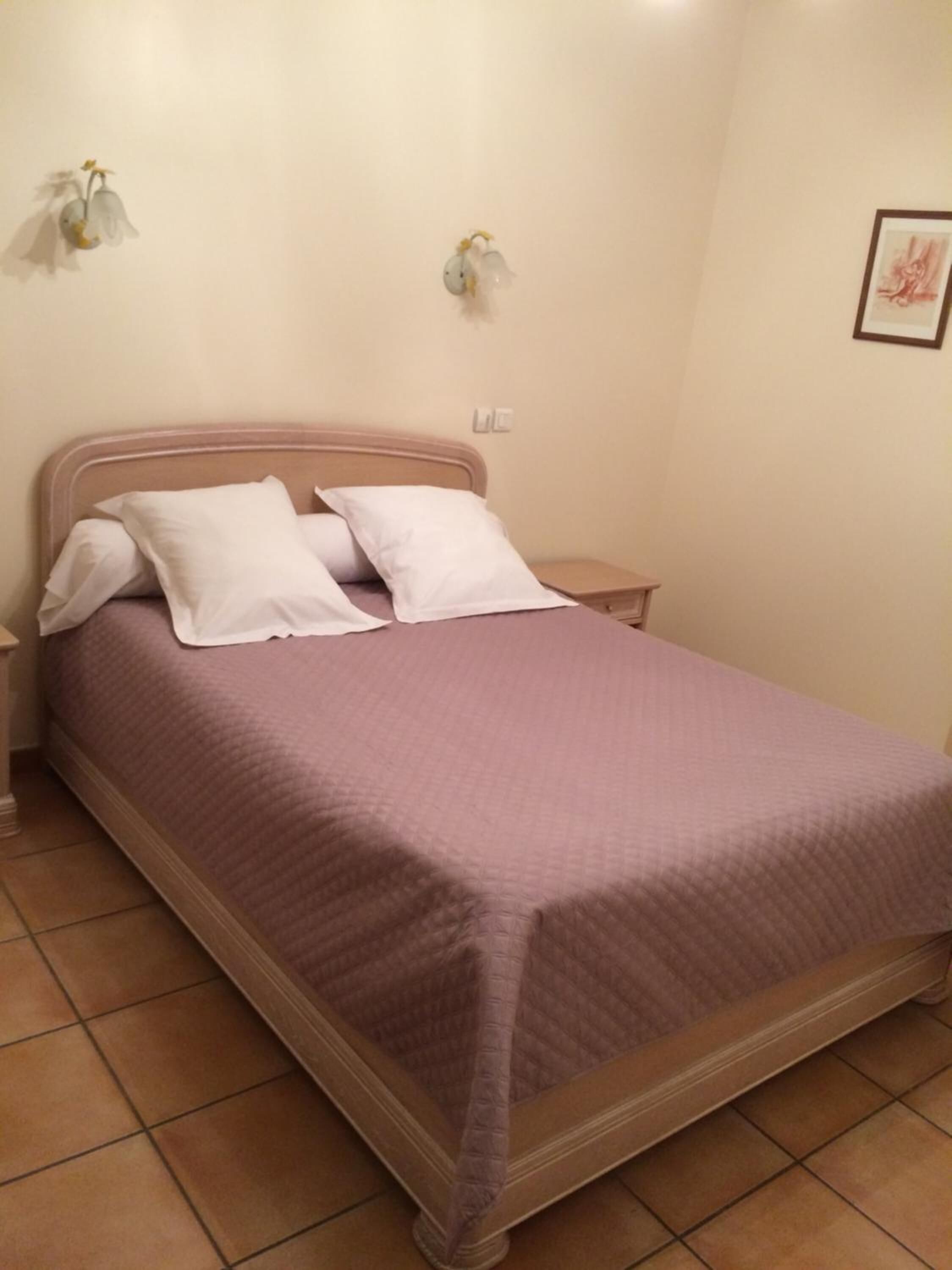 Un lit simple avec un cadre en bois, une literie blanche et une couvre-lit de couleur violet pâle. Il y a deux tables de chevet avec des lampes, et un tableau encadré accroché au mur.