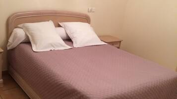 Un lit simple avec un cadre en bois, une literie blanche et une couvre-lit de couleur violet pâle. Il y a deux tables de chevet avec des lampes, et un tableau encadré accroché au mur.