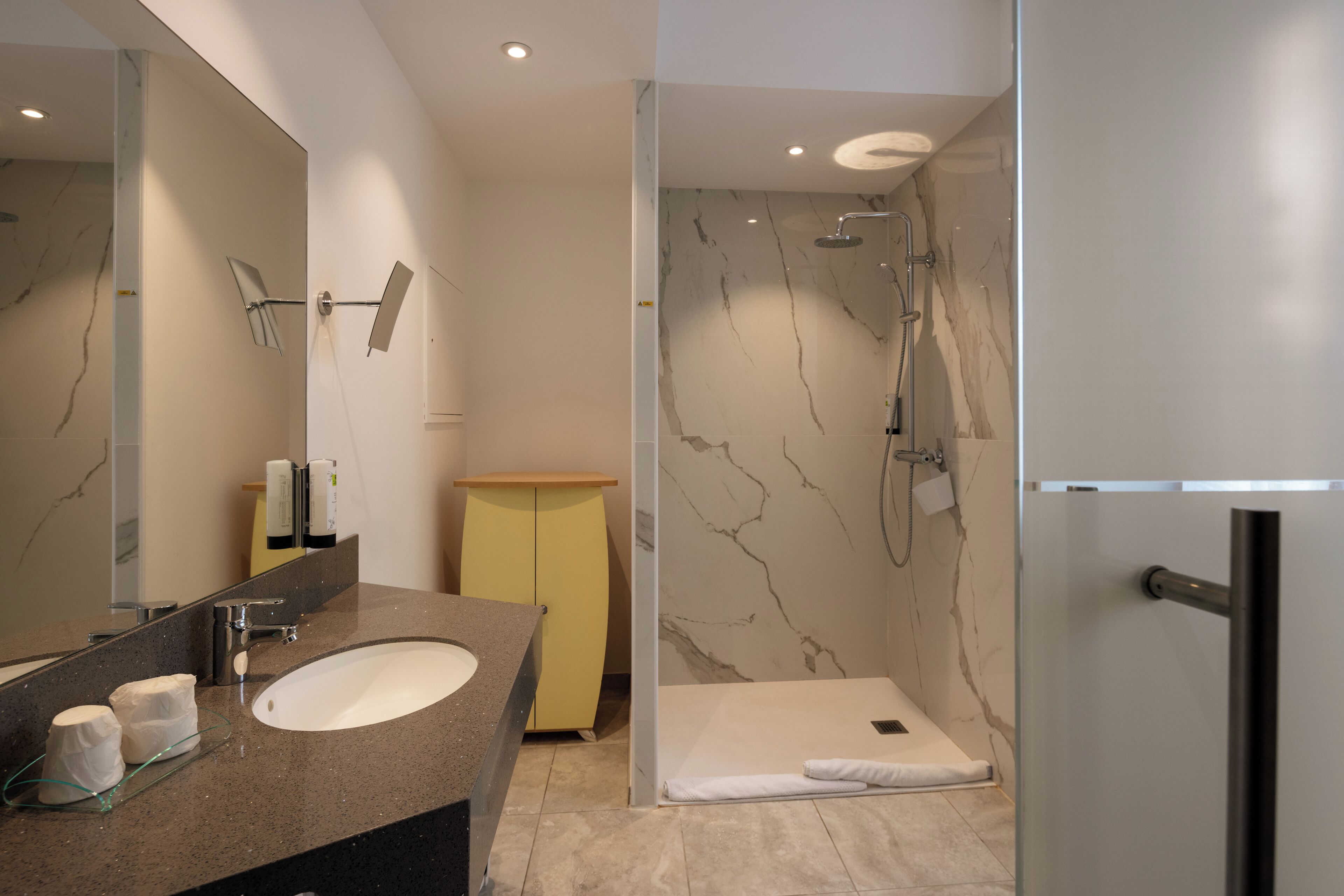 A modern bathroom with a marble shower, a sink with a glass tray, and a yellow cabinet.