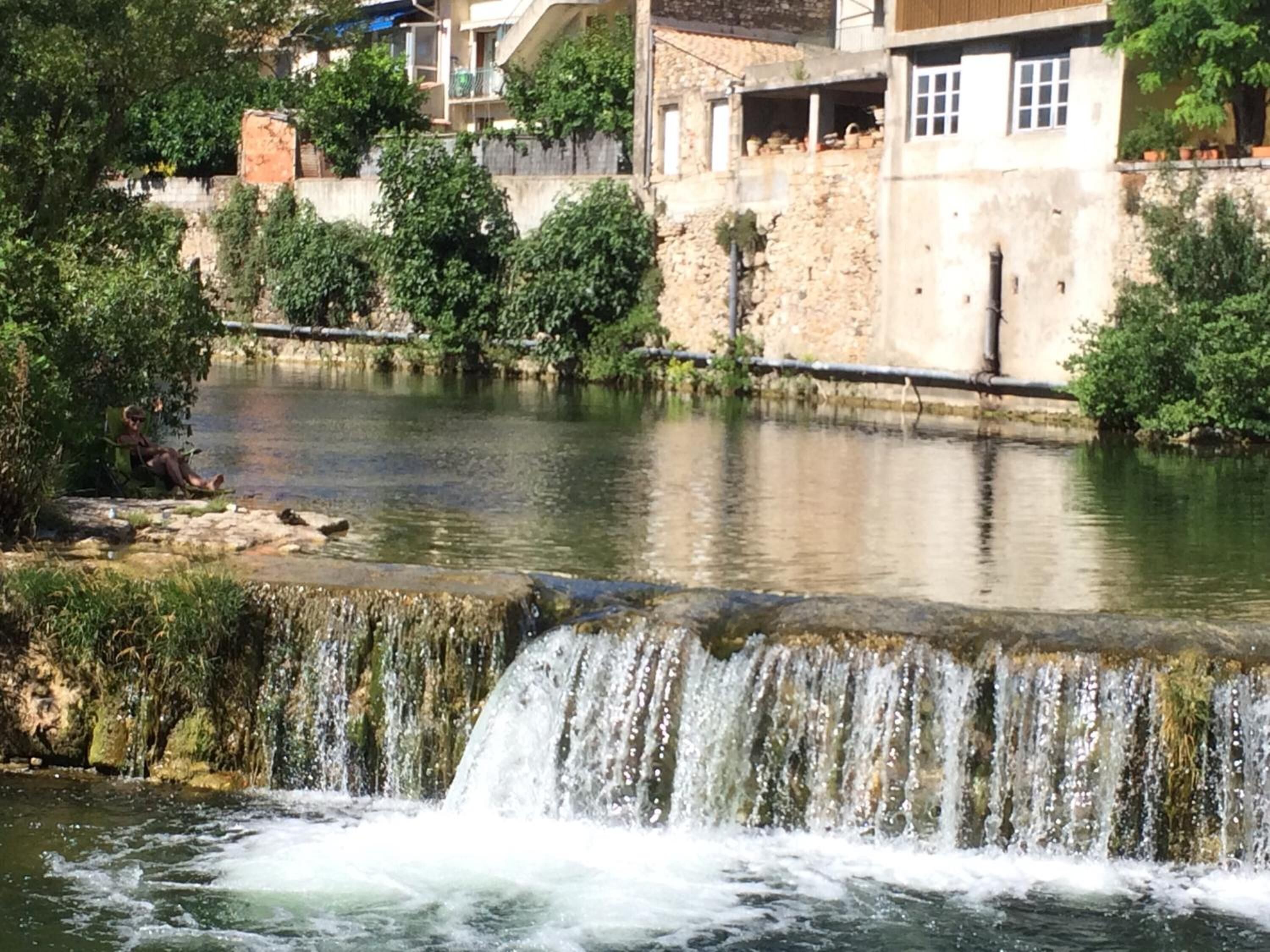A waterfall in a natural setting with a stone wall and greenery in the background.
