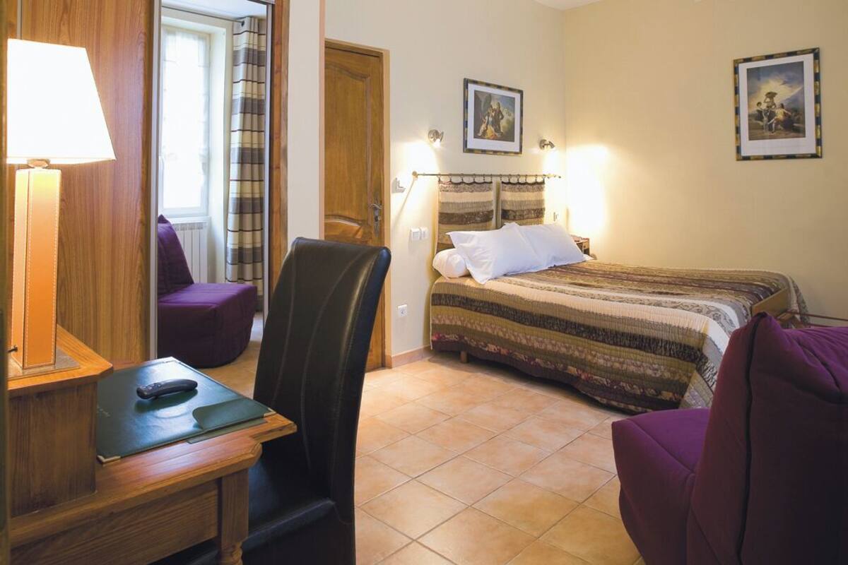 A hotel room with a bed, a desk with a lamp, a chair, and a window with curtains.