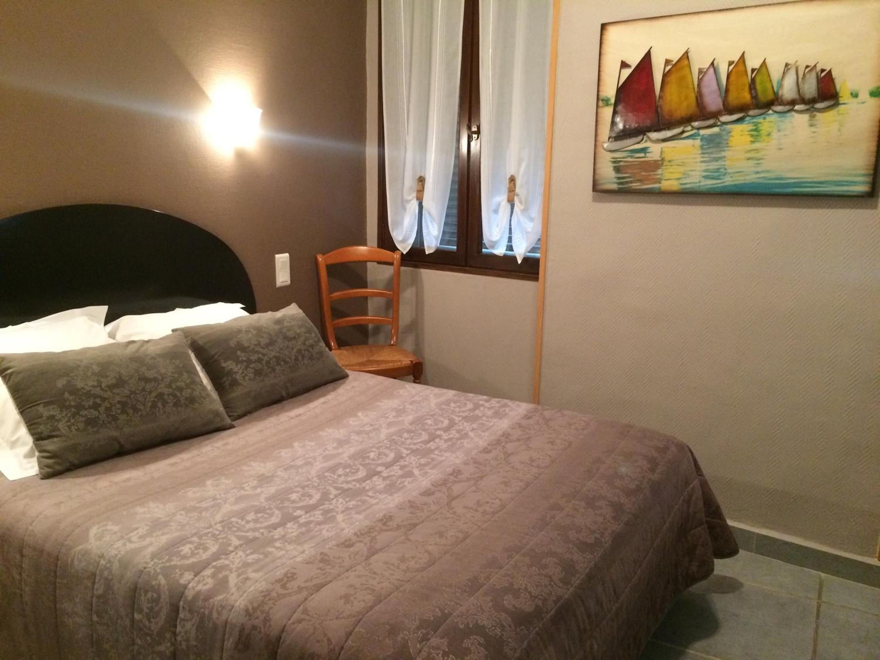 Un lit bien fait, recouvert d’une couvre-lit à motifs, une chaise en bois et un tableau représentant des voiliers accroché au mur.