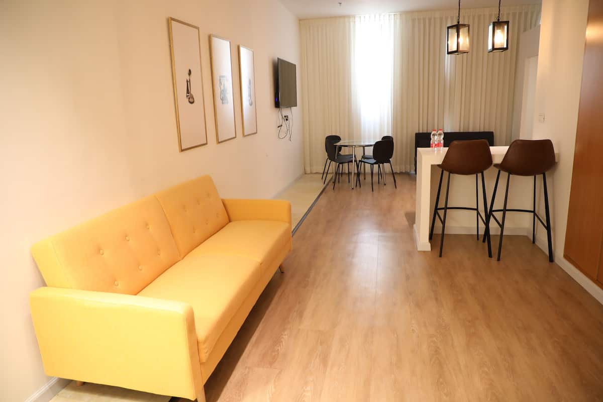 A modern living room with a yellow sofa, a dining area with a table and chairs, and a TV mounted on the wall.
