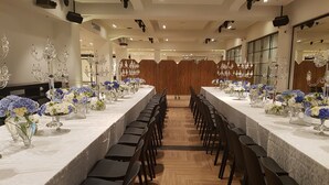 Una larga mesa de banquete con sillas, decorada con centros de mesa florales, en una sala bien iluminada con suelo de madera y techo alto.