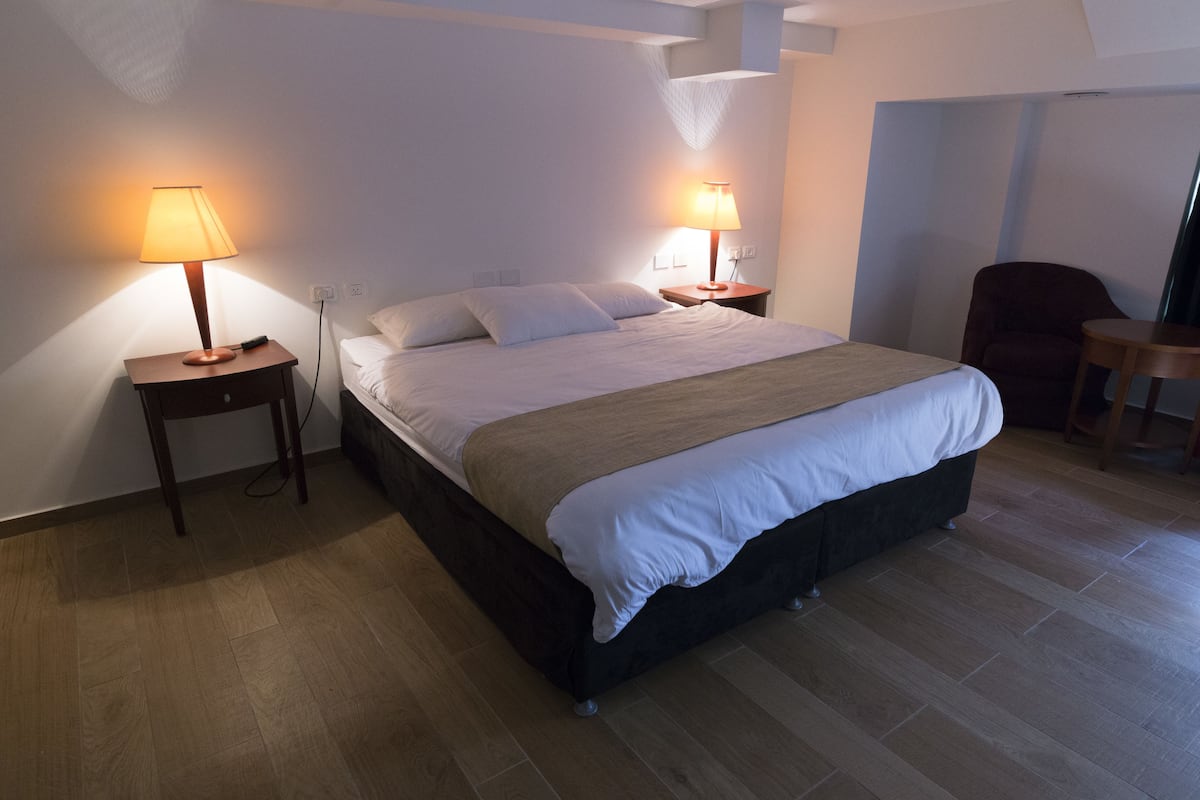 A hotel room with a large bed, two bedside tables with lamps, a wooden floor, and a chair in the corner.