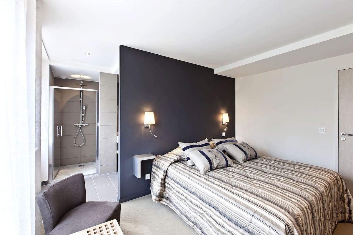 Habitación de hotel moderna con cama, lámparas de noche, una silla y zona de ducha.