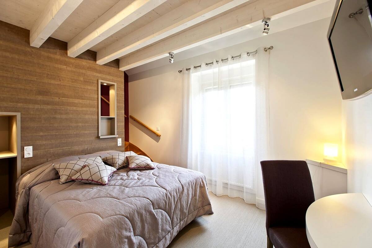 Un dormitorio con cama, una silla, un televisor, un ventanal con cortinas y techo de madera.