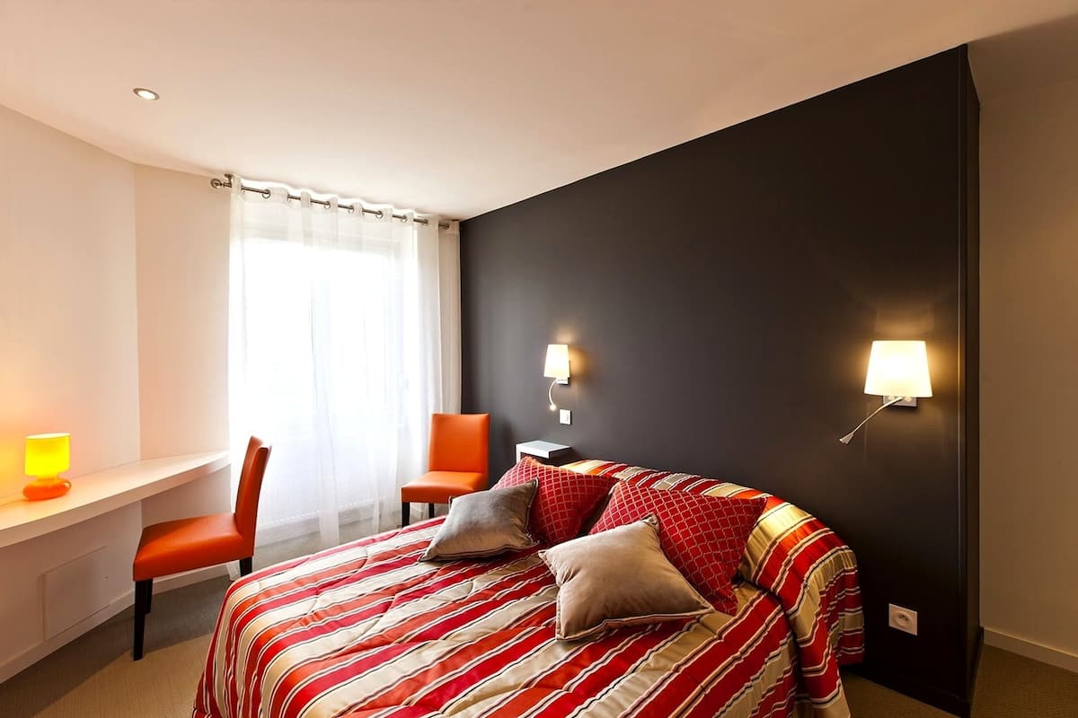 Habitación de hotel con cama, escritorio, silla y dos lámparas fijadas a la pared.