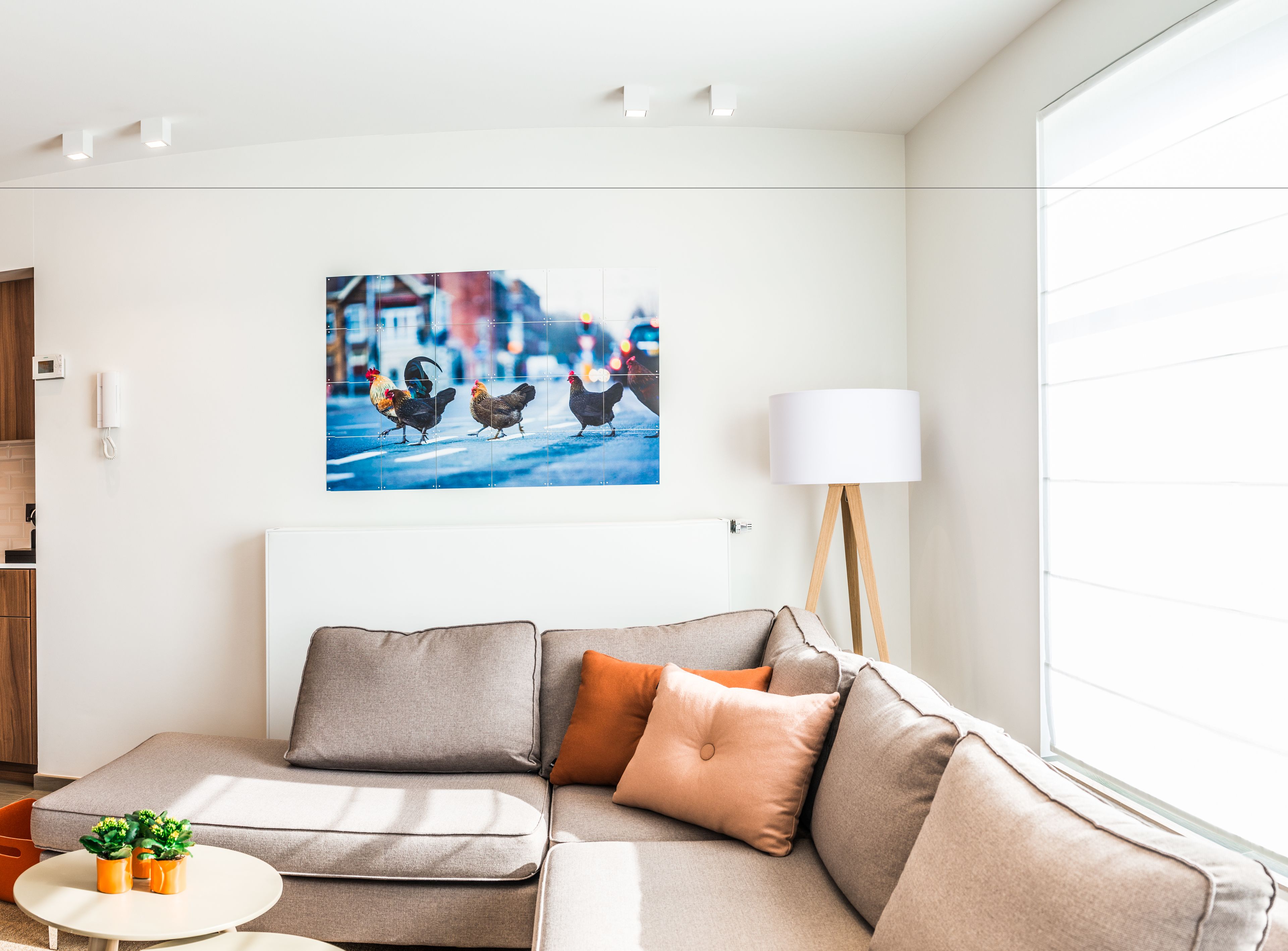 Un salon moderne avec un canapé gris, des coussins orange, une lampe blanche et un tableau représentant un paysage urbain accroché au mur.