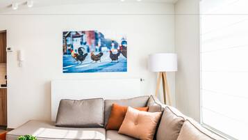 Un salon moderne avec un canapé gris, des coussins orange, une lampe blanche et un tableau représentant un paysage urbain accroché au mur.