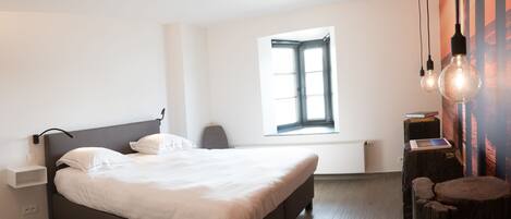 A modern bedroom with a large bed, a bedside table, a chair, a window with blinds, and a wall-mounted light fixture.