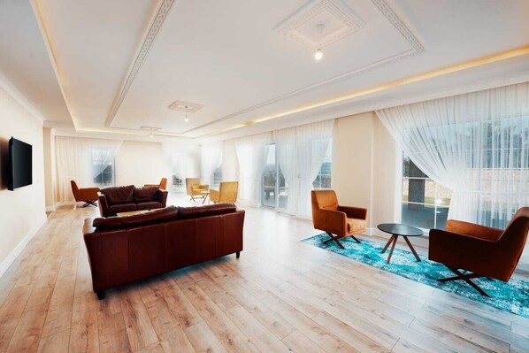 A spacious living room with a large brown sectional sofa, two armchairs, a coffee table, and a flat-screen TV mounted on the wall.
