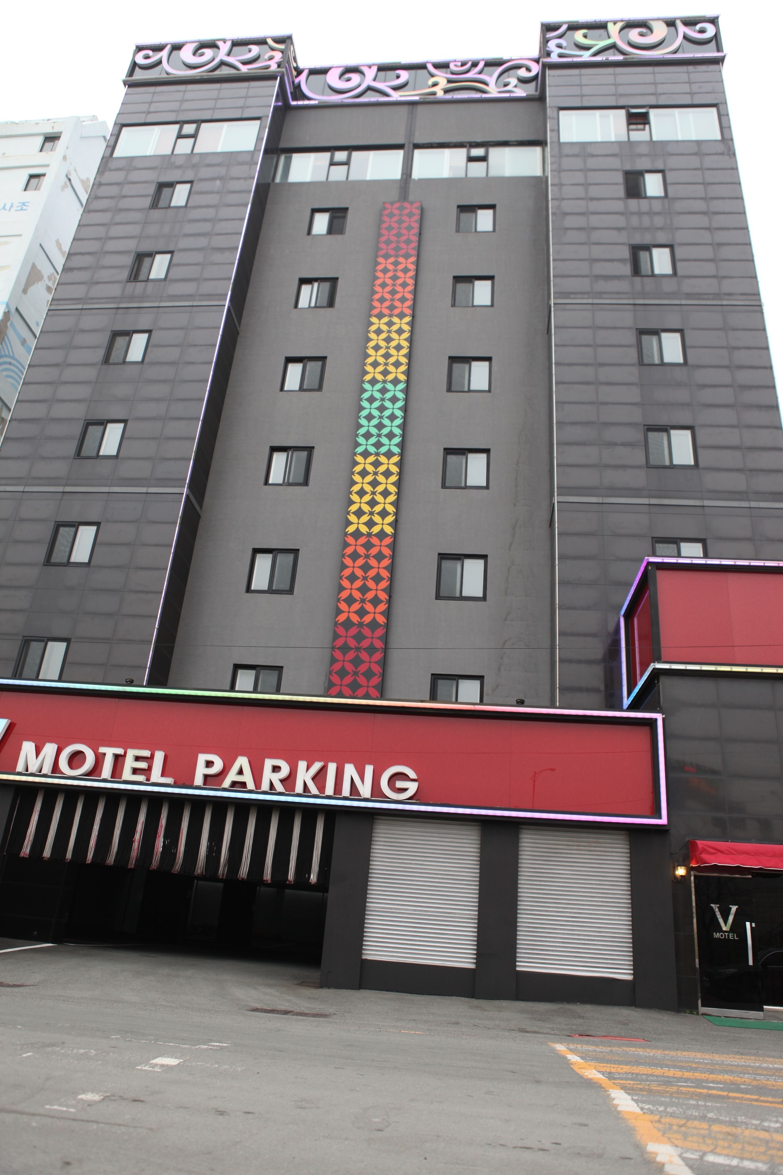 A multi-story building with a vertical stripe pattern on the facade, a red sign reading "Motel Parking," and a visible entrance with a closed shutter.