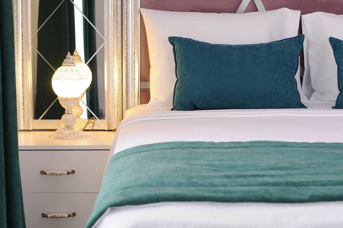 Un lit bien fait, avec une literie blanche et turquoise, une lampe de chevet et un miroir doté d’un cadre décoratif.