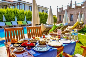 A dining table set outdoors with various dishes, surrounded by lounge chairs and umbrellas, near a pool and a historic building. - Cunda Rota Hotel (Ayvalik)