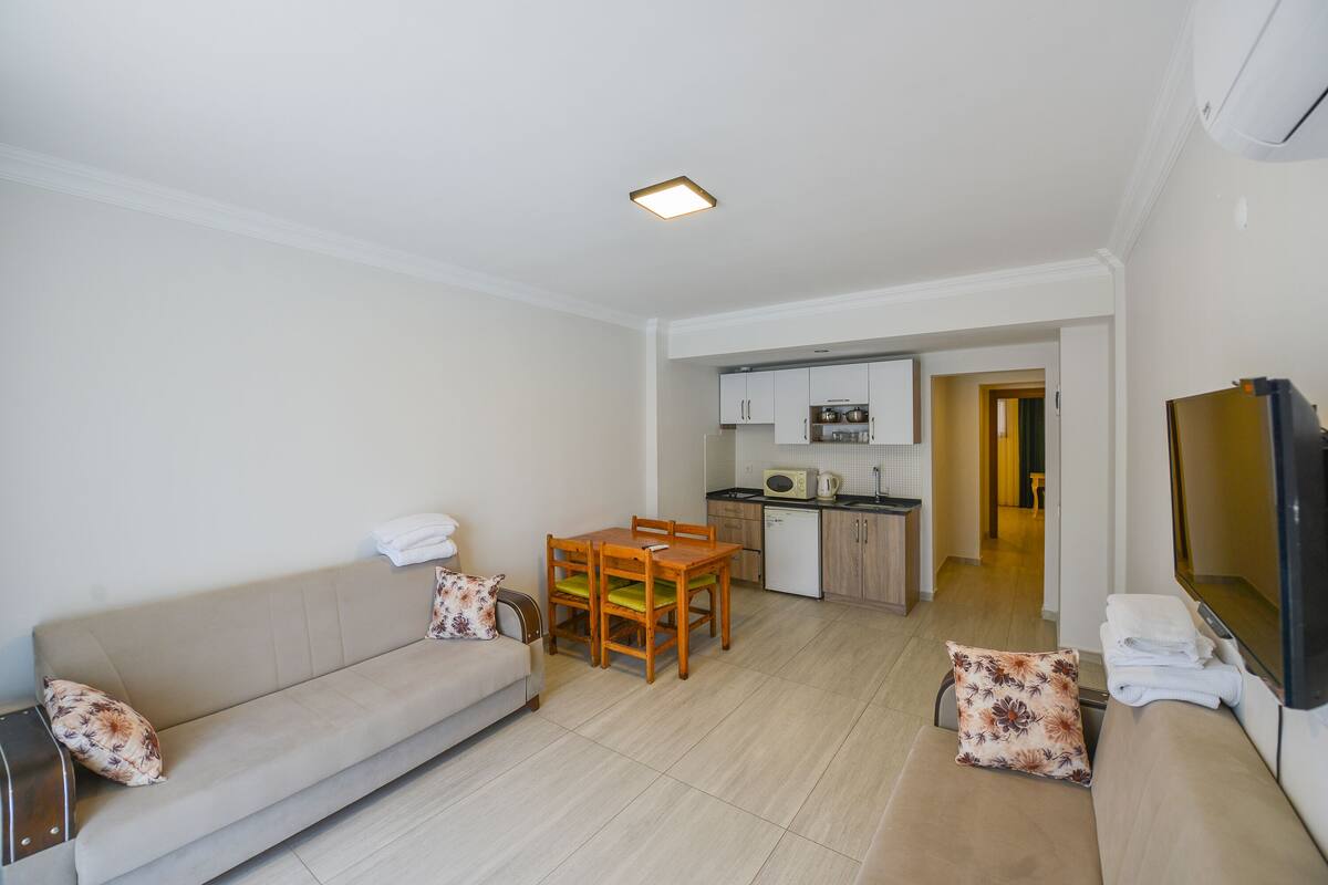 A living room with a sofa, a dining table, and chairs. There is a kitchen area with cabinets and a microwave. A TV is mounted on the wall.