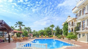 A swimming pool with lounge chairs and umbrellas, surrounded by palm trees and a building with balconies. - Milkyway Apart & Hotels (Fethiye)