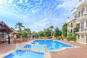 Piscina con sillones de descanso y sombrillas, rodeada de palmeras y un edificio con balcones.