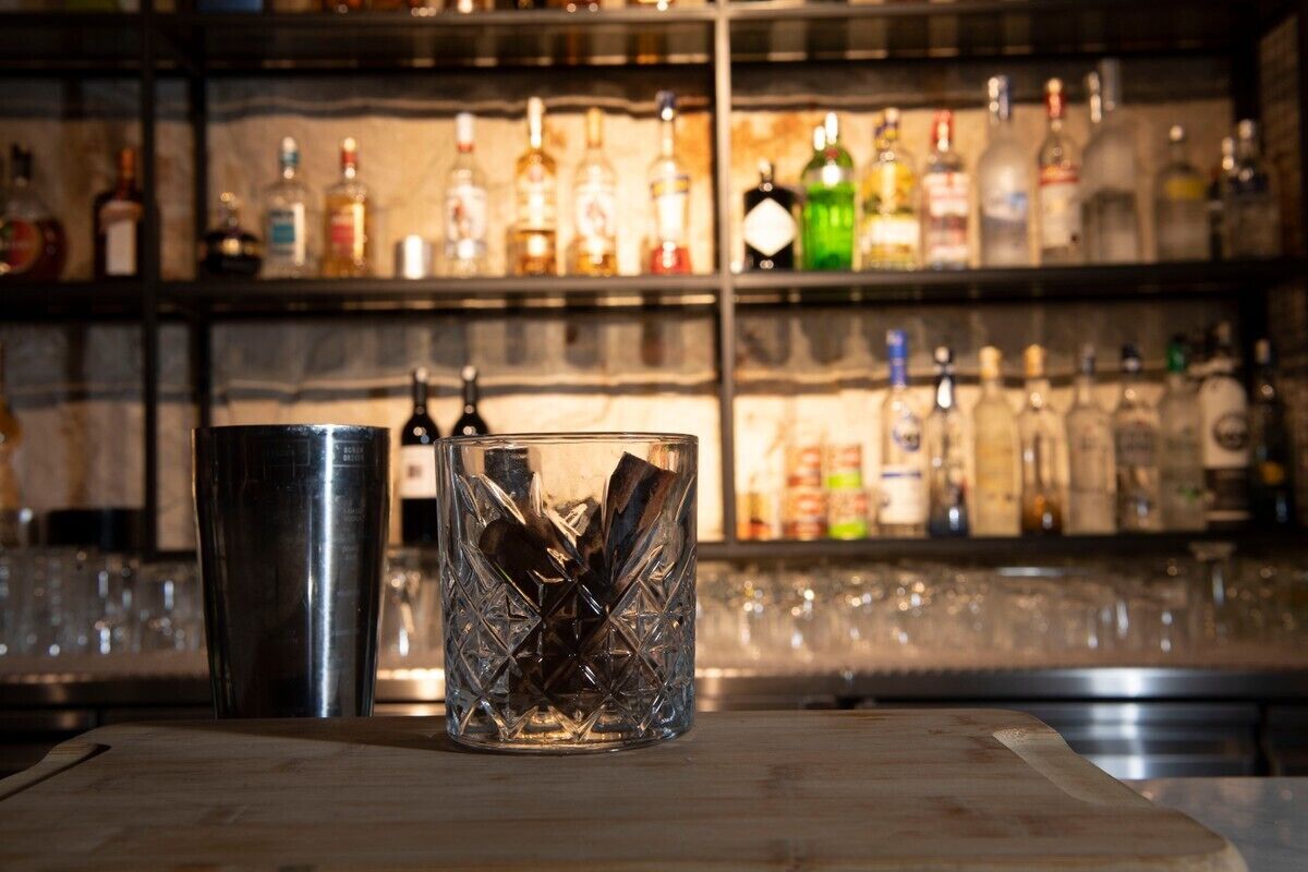 Uma área de bar com um recipiente de vidro e um coqueteleira de metal sobre uma superfície de madeira, com prateleiras cheias de várias garrafas ao fundo.