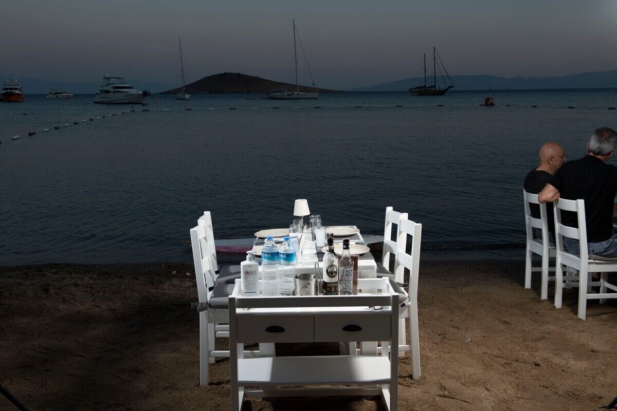 A table set up on a sandy beach with a view of the sea and boats in the background.
