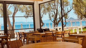 Een restaurant met houten meubels en grote ramen, met uitzicht op zee en bomen.