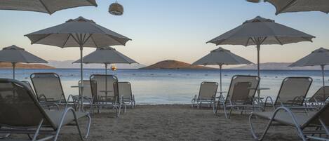 A beach with rows of sun loungers and umbrellas, a boat on the water, and a hilly landscape in the distance.