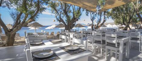 An outdoor dining area with white tables and chairs, arranged under large umbrellas, overlooking a beach with umbrellas and chairs.
