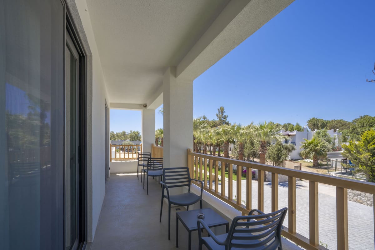 A balcony with chairs and tables, overlooking a street with palm trees and buildings.