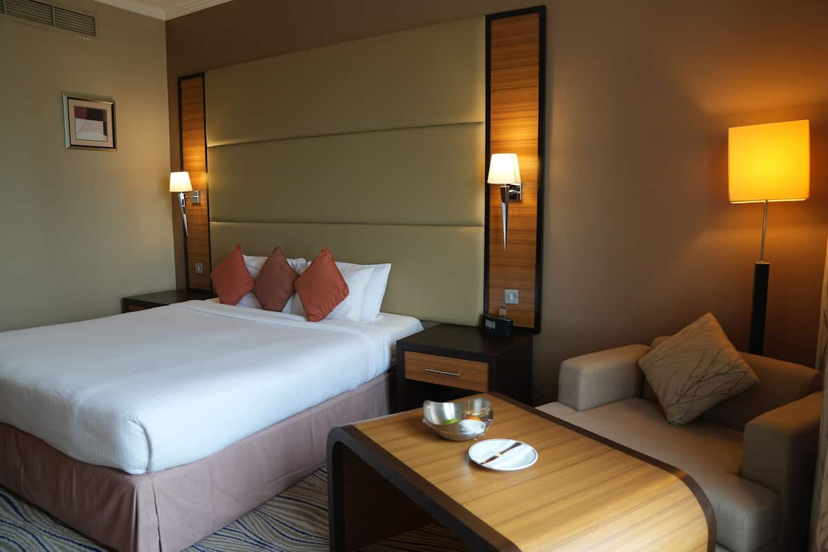 Una habitación de hotel con una cama grande, una mesita de noche con lámpara, una pequeña mesa con un cuenco y un plato, y un sofá.