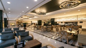 Eine geräumige Lobby mit Schachbrettmuster-Boden, mehreren Sitzbereichen inklusive Sofas und Sesseln, einer Bar und einer Treppe zu den oberen Stockwerken.