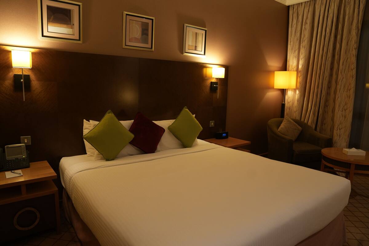 Habitación de hotel con una cama grande, dos mesitas de noche, una silla y una lámpara.