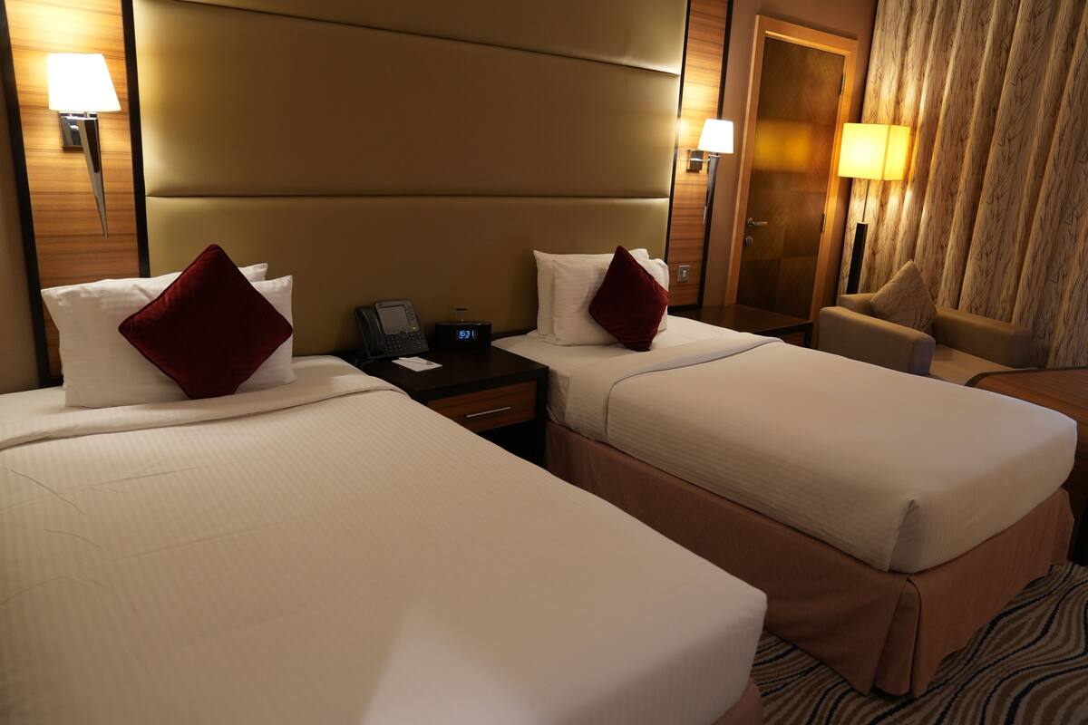 Habitación de hotel con dos camas, cada una con ropa blanca y almohadas rojas, una mesita de noche con teléfono y reloj digital, y una silla con lámpara.
