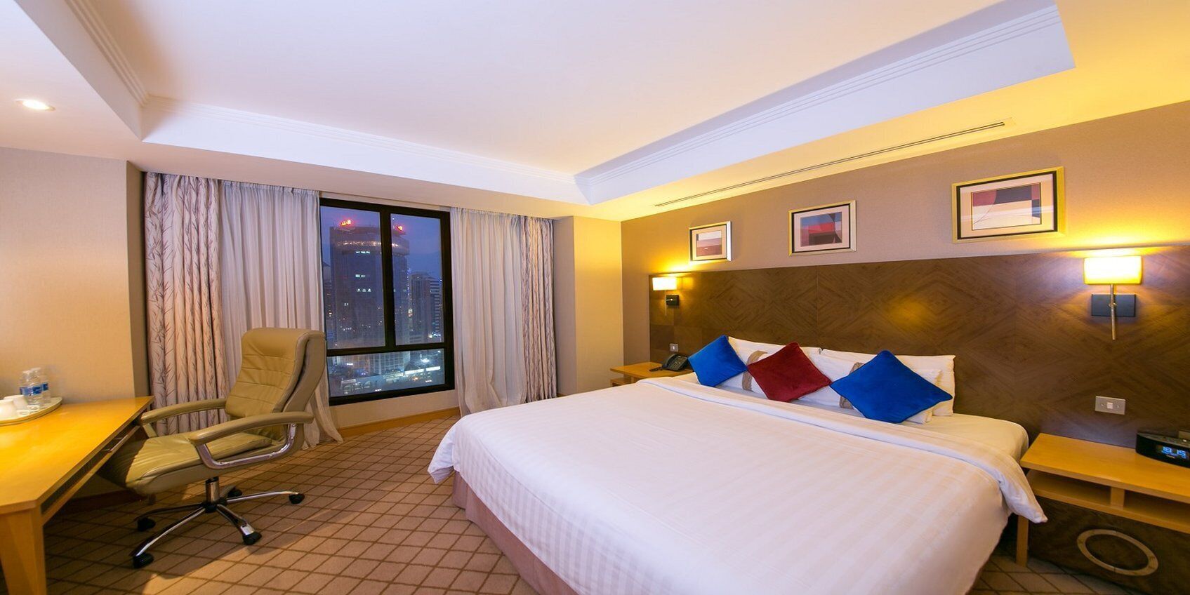 A hotel room with a large bed, a desk, a chair, and a view of a cityscape through a window.