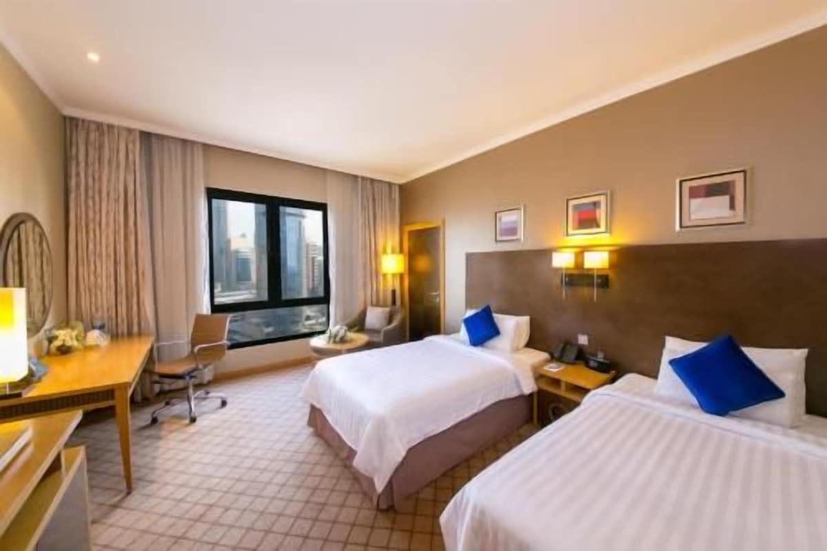 A hotel room with two beds, a desk, a chair, a window with a view, and wall-mounted lights.