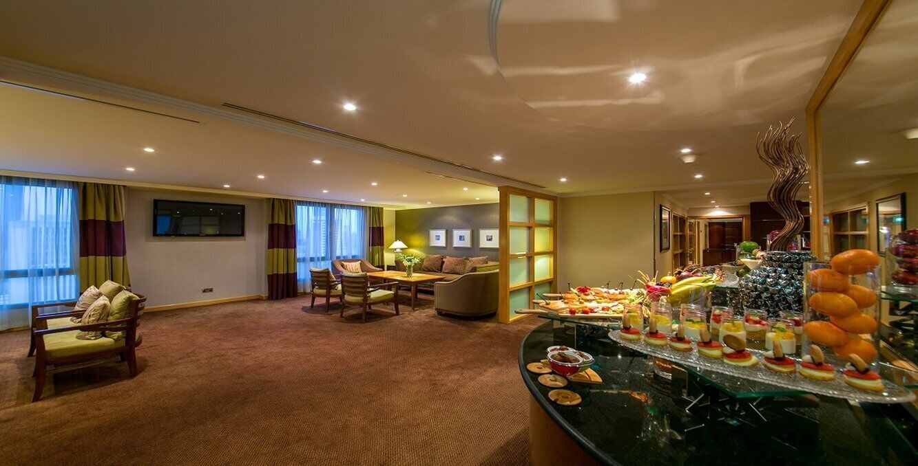 A hotel room with a dining area, a sofa, a table with a lamp, and a buffet with various desserts and fruits.