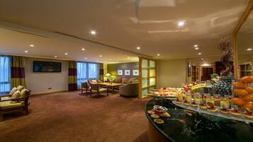 A hotel room with a dining area, a sofa, a table with a lamp, and a buffet with various desserts and fruits.