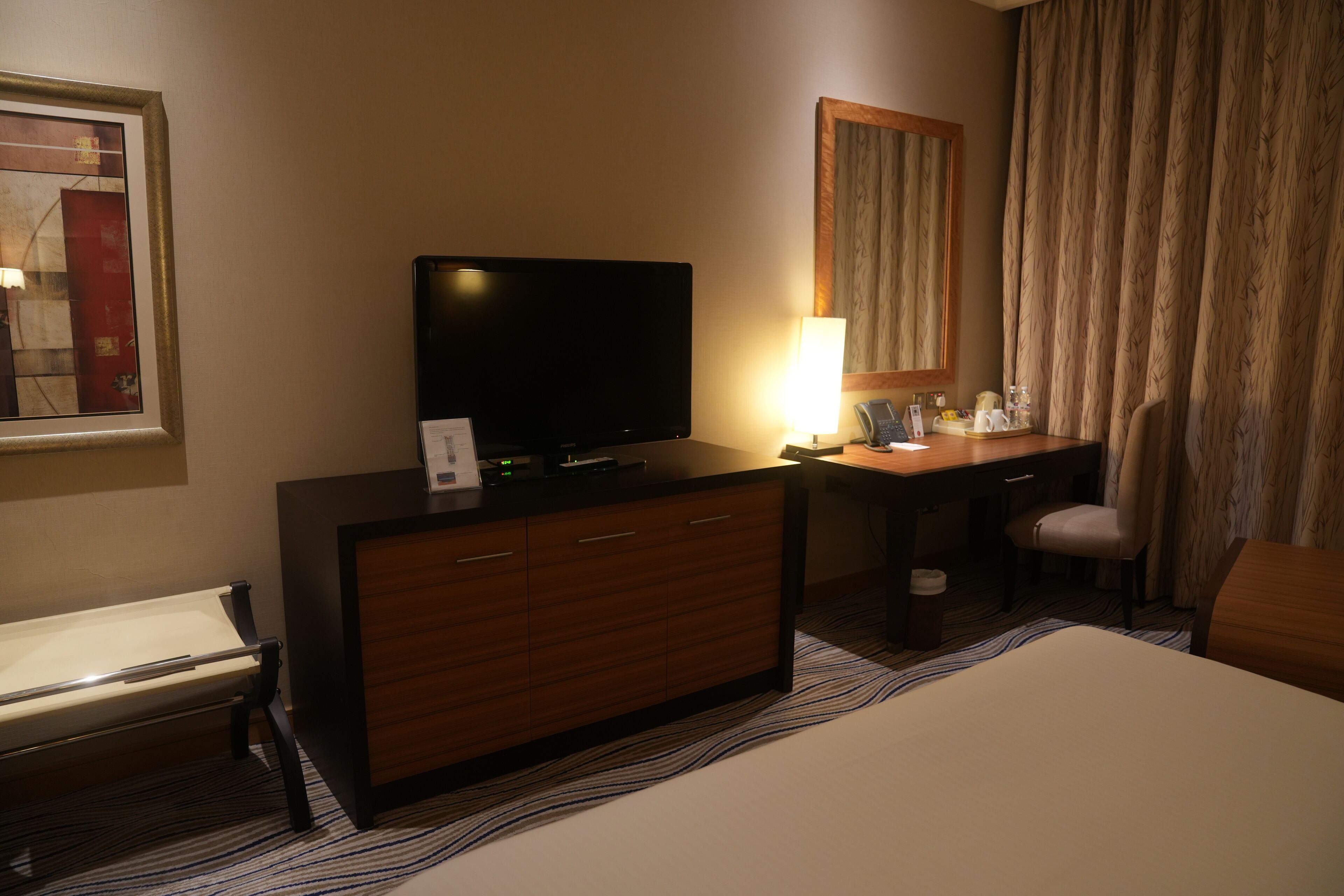 A hotel room with a bed, a desk, a television, a lamp, a chair, and a large window with curtains.