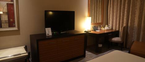 A hotel room with a bed, a desk, a television, a lamp, a chair, and a large window with curtains.