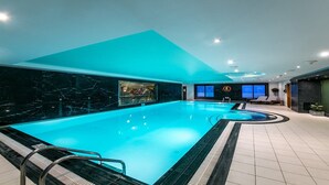 Ein großer, überdachter Pool mit blauer Beleuchtung, eine Lounge mit Sesseln und Blick auf die Stadt durch die Fenster.