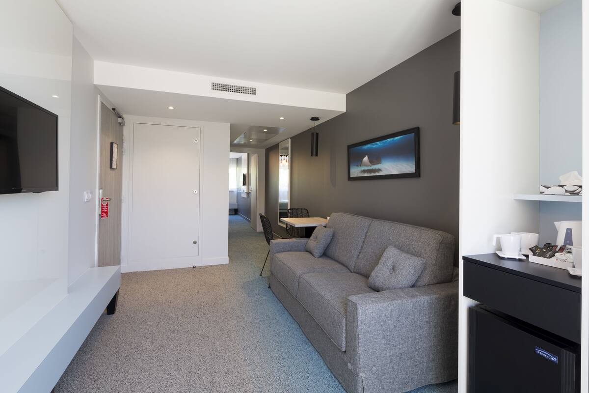 Une chambre d’hôtel moderne avec un canapé gris, un téléviseur à écran plat, une petite kitchenette et un coin repas comprenant une table et des chaises.