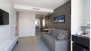 Una habitación de hotel moderna con un sofá gris, un televisor de pantalla plana, una kitchenette pequeña y un comedor con mesa y sillas.