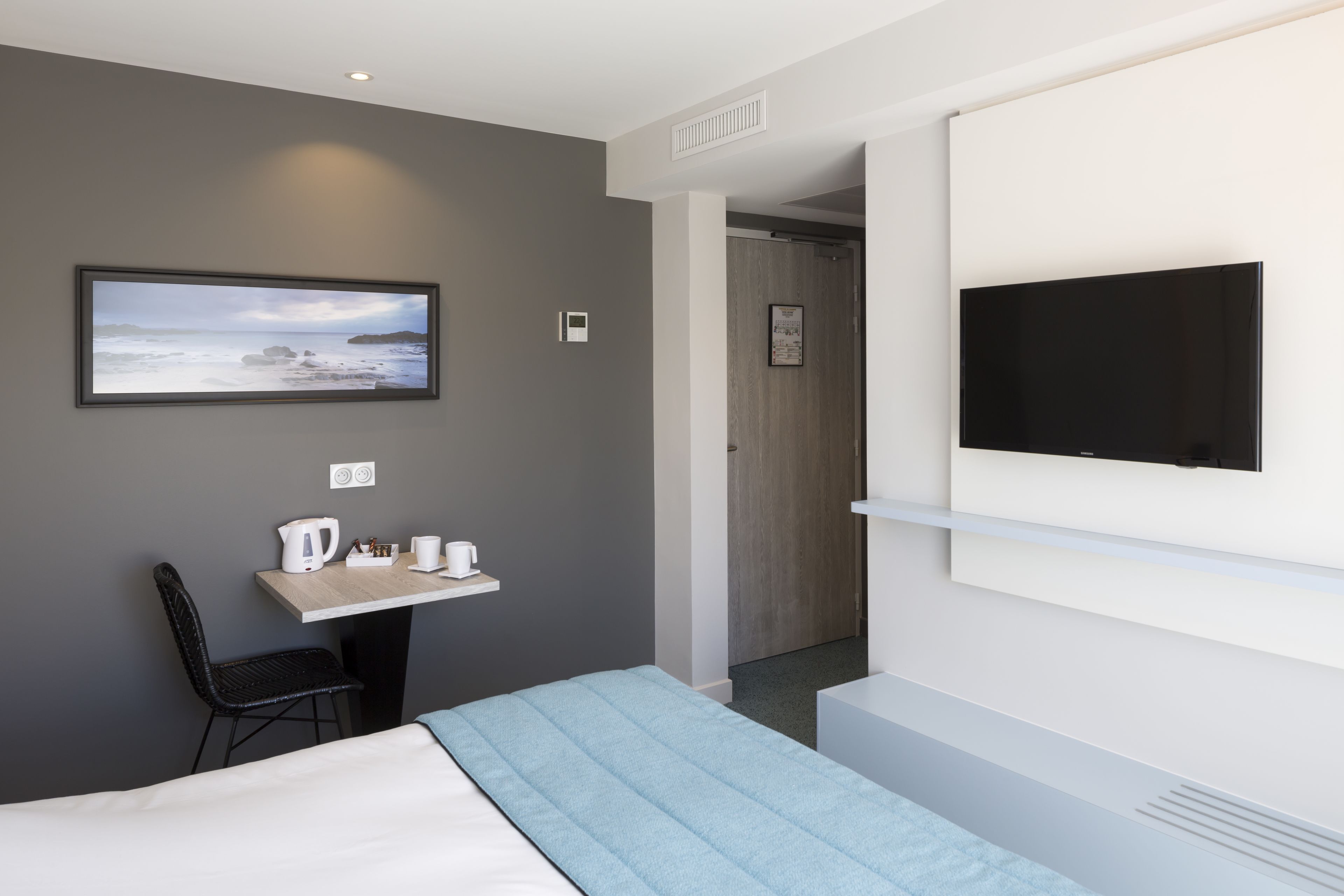 A modern hotel room with a bed, a desk with a kettle, a chair, a flat-screen TV mounted on the wall, and a framed picture of a beach scene.