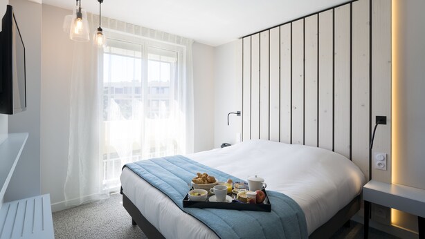 Una habitación de hotel moderna con una cama grande, una mesita de noche, una bandeja con alimentos para el desayuno y una ventana con cortinas traslúcidas.