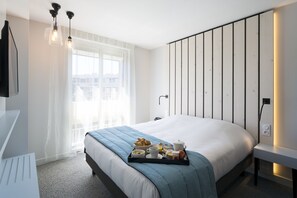 A modern hotel room with a large bed, a bedside table, a tray with breakfast items, and a window with sheer curtains. - Opéralia Hôtel Les Pins (Balaruc-les-Bains)